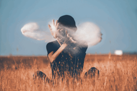Image: a man in black clothing sits in a field of amber grain against a blue sky. He has thrown two fistfuls of sand into the air which obscure his face and appear almost like clouds of smoke.