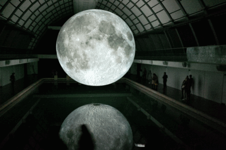Image: photo of a darkened interior gallery in which people observe Luke Jerram's art installation titled Museum of the Moon, a highly-detailed and illuminated scaled replica of the moon which hovers in mid-air.