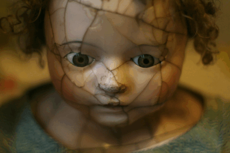 Image: the vacant stare of an antique porcelain doll, the surface of which is splintered with cracks.