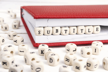 Image: a red hard-covered book lies on a table, propped slightly open in the middle by small wooden blocks with letters printed on their faces, spelling the word "subtitle".