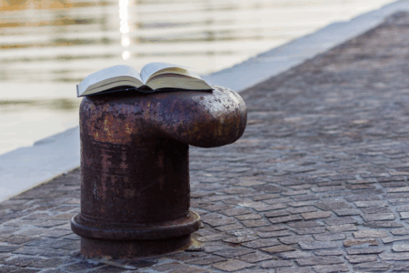 Image: a book lies open atop a mooring bollard near the edge of an urban waterway.