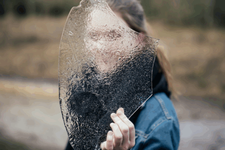 Image: a woman holds a shard of frosted glass in front of her, obscuring the view of her face.