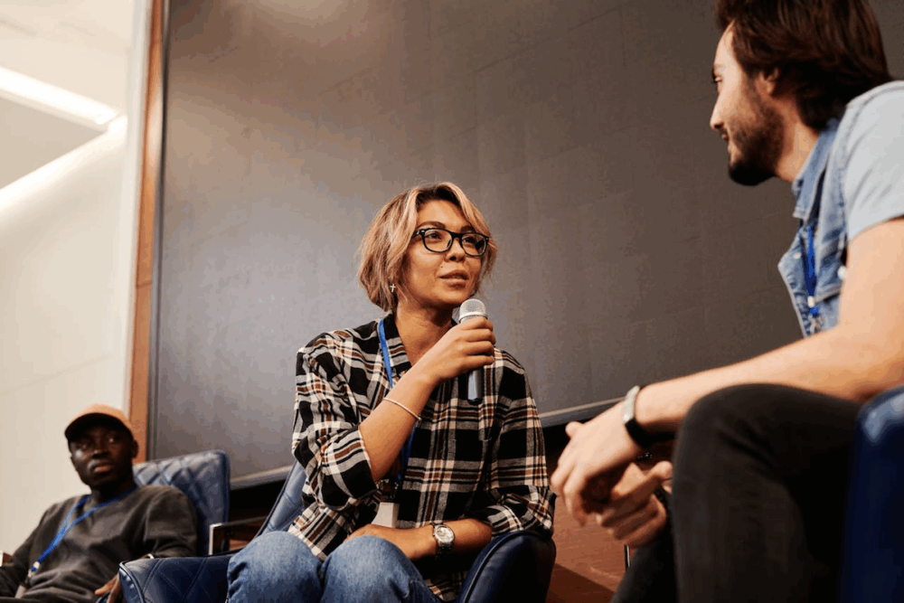 Image: at a panel discussion on a convention stage, the female moderator holding a microphone asks a question to a male panelist.