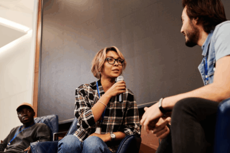 Image: at a panel discussion on a convention stage, the female moderator holding a microphone asks a question to a male panelist.