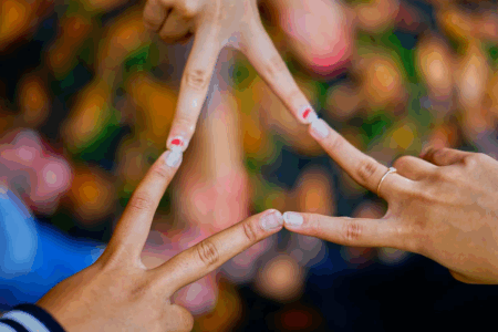 Image: three women's hands, each one extending their index and middle fingers in a V shape, the fingertips touching so that the three V's form a continuous triangle.