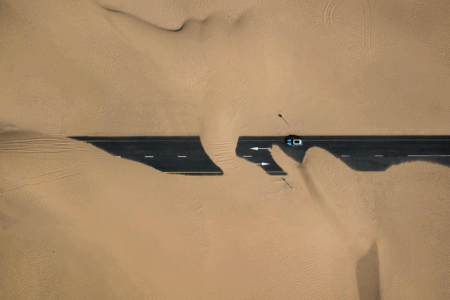 Image: aerial view of a two-lane road cutting through the desert and which has been completely obscured by drifting sands, leaving a car unable to proceed.