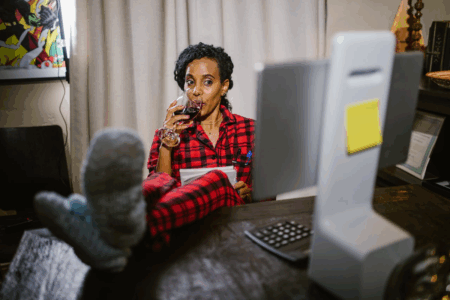 Image: a woman wearing pyjamas and cozy socks sits with her feet up on her desk, holding a notebook in her lap and drinking a glass of red wine while watching something on her computer's screen.