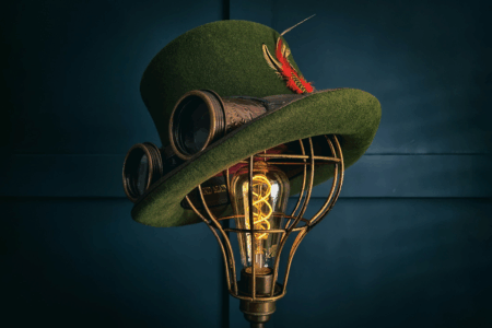 Image: a dark green felted top hat accented with small colorful feathers and outfitted with steampunk-style eye goggles sits atop a bulb-shaped wire cage that holds a dimly-lit antique lightbulb.
