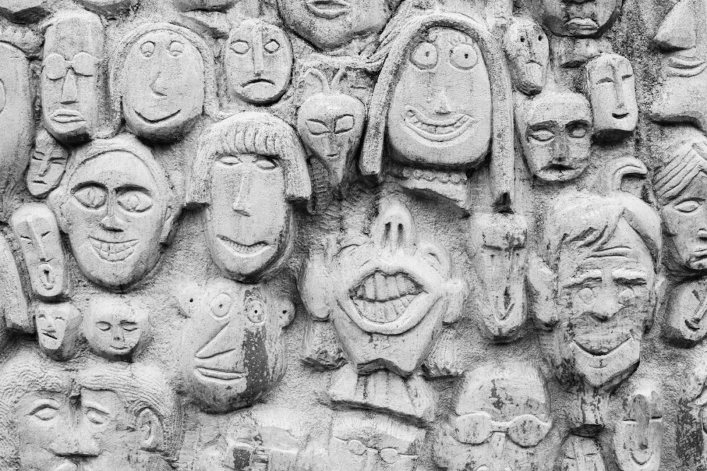 Image: a concrete wall covered with dozens of unique faces sculpted in three-dimensional relief.