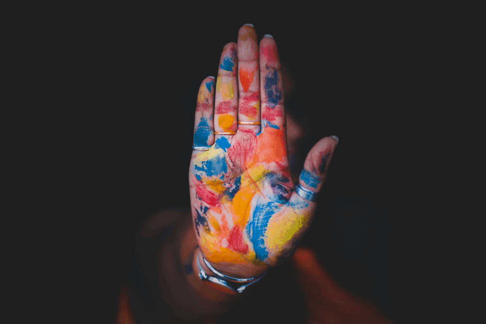 a woman's hand, the palm and fingers of which are daubed with many colors of paint, is held up to the viewer as if to signal 'stop'.