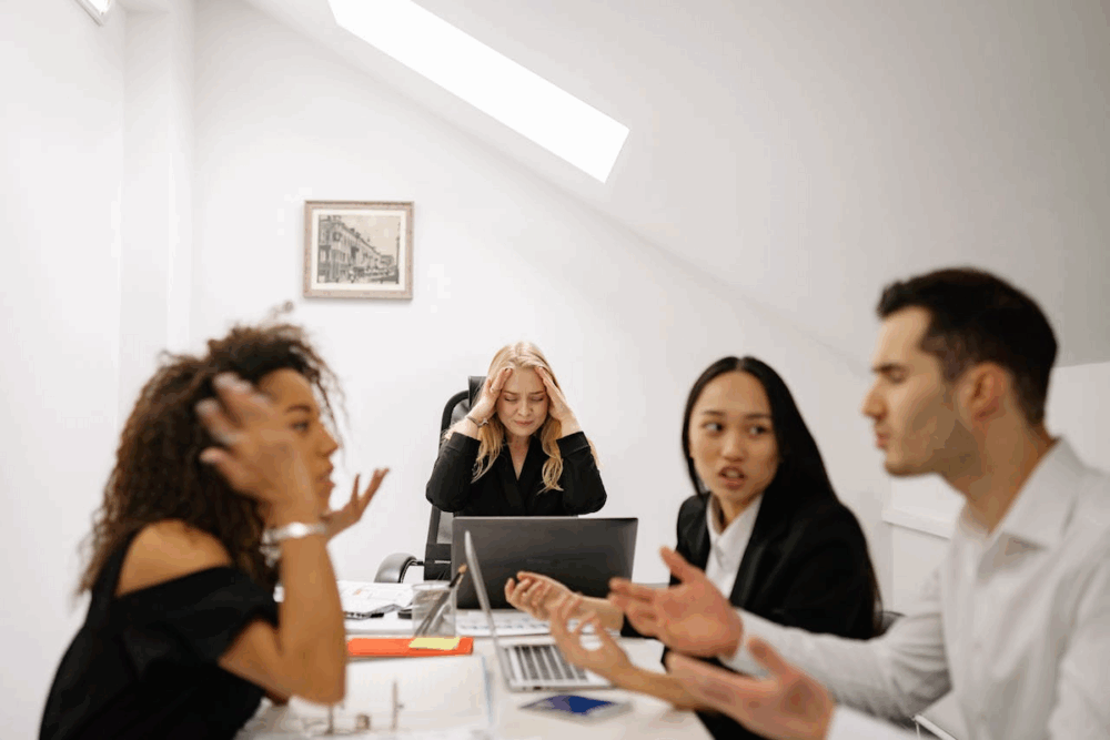 Image: while three colleagues engage in heated discussion, an office worker holds her hands to her anguished face.