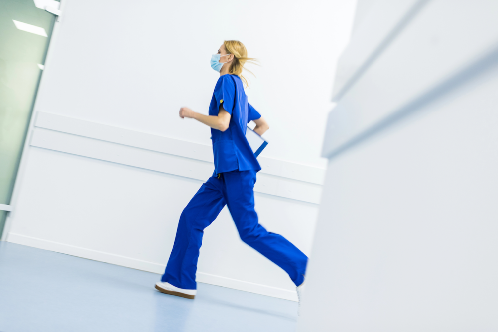 Image: a nurse in blue scrubs runs down a hospital corridor.