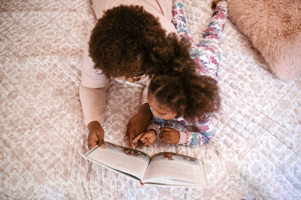 Image: a mother and daughter lie across a bed, reading a book together.