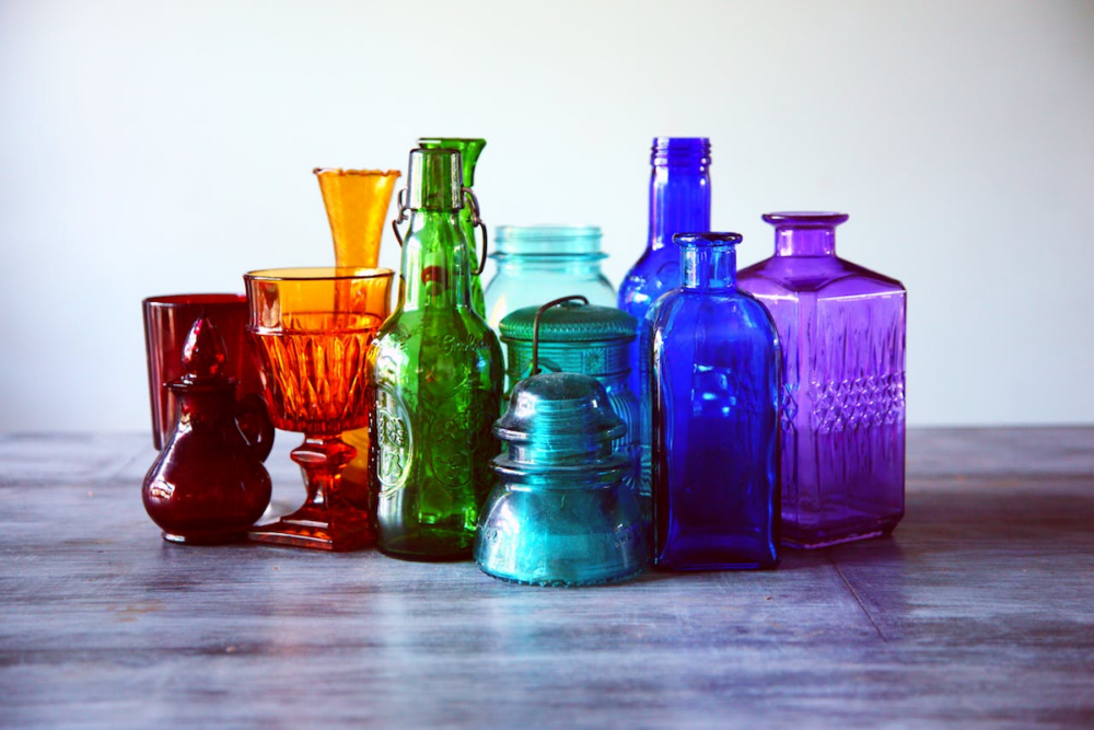 Image: an assortment of colored glass carafes, glasses, bottles and jars representing the full chromatic spectrum stand together on a tabletop.