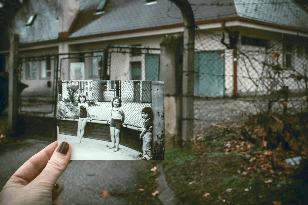 Image: in front of a dilapidated building's chain link fence a woman's hand holds a black and white photo of children standing in the same spot decades earlier.