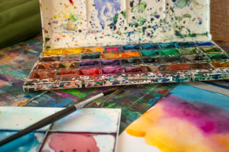 Image: an open tray of watercolor paints and a brush sit next to a painting in progress.
