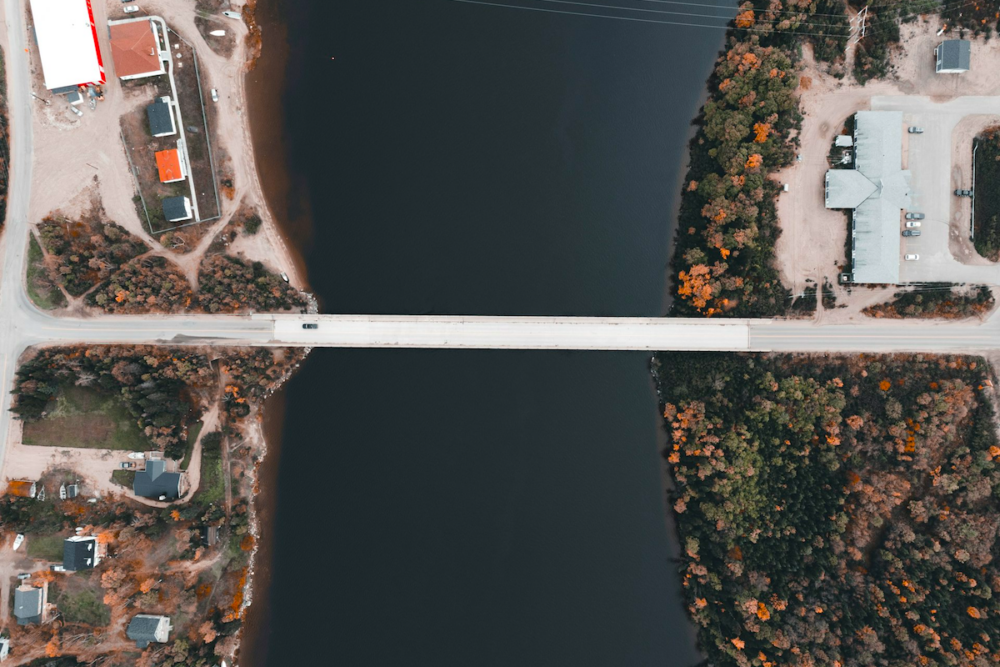 Image: seen from an aerial view, a single car drives across a bridge that connects two sides of town across a waterway.