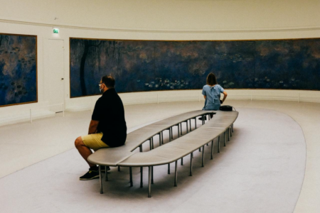 Image: Two museum-goers sit on a bench in the center of a round gallery looking at long Monet waterlily paintings mounted on the walls.