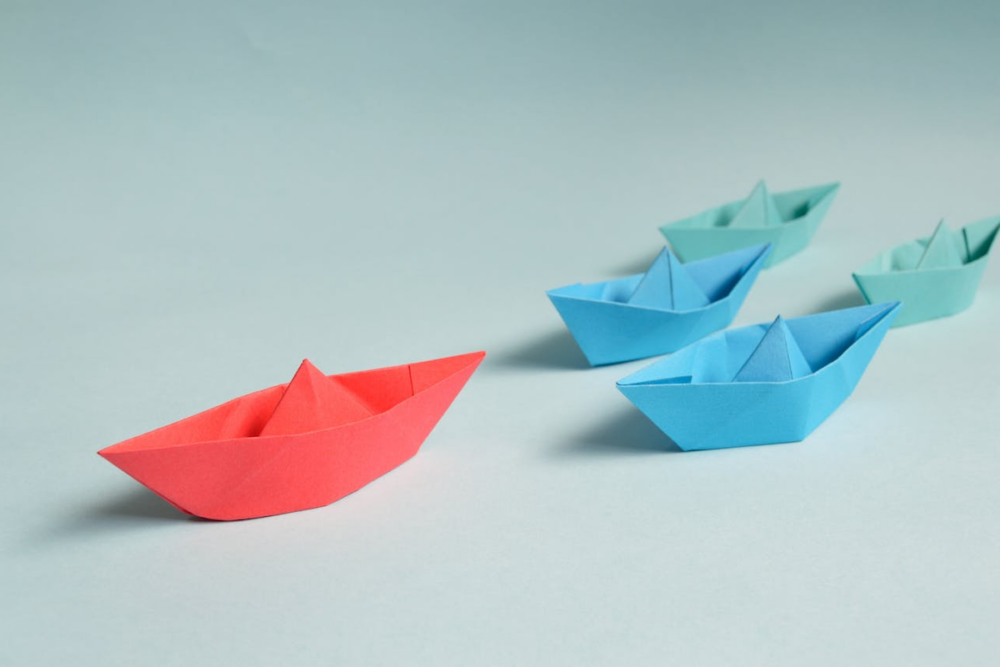 Image: Five origami boats are arranged on a tabletop: the largest red boat in front, followed by two smaller blue boats, followed by two smaller green boats.