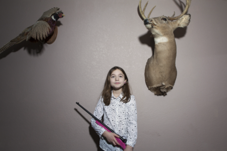 Image: a young girl holding a child's-size hot pink hunting rifle stands in front of a wall on which are mounted a taxidermied ring-tailed pheasant and the head of an eight-point buck.