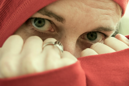 Image: close-up photo of a woman staring straight at the viewer with her hoodie pulled over her head and balled fists held in front of her face.