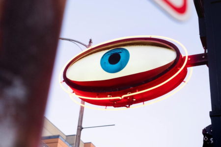 Image: An oval-shaped sign painted to look like an open eye, encircled with neon lights, extends from the face of an urban building.