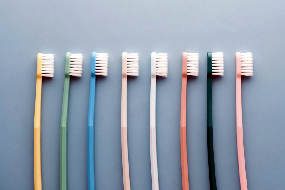 Image: eight inexpensive toothbrushes of different colors lie on their sides in a row on a blue backdrop.
