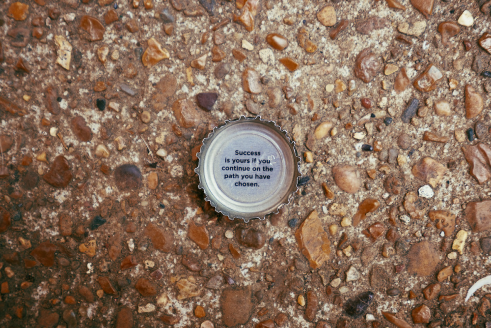 Image: a bottle cap with the words "Success is yours if you continue on the path you have chosen" printed on the inside lies on the ground facing upward.