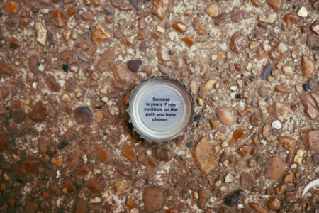 Image: a bottle cap with the words "Success is yours if you continue on the path you have chosen" printed on the inside lies on the ground facing upward.