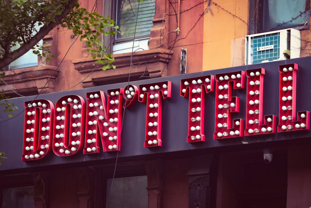 Image: The words "Don't Tell" in an illuminated marquee mounted across building facades on an urban street.