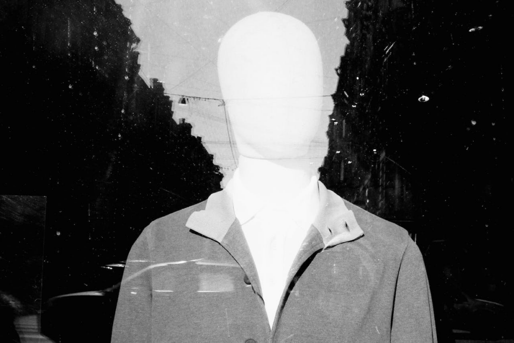 Image: a blank-faced mannequin is seen through the reflections on a shop window.