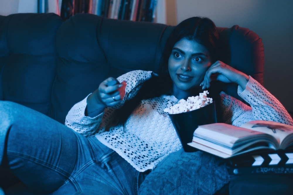 Image: a young woman lies on her sofa watching television, an open book beside her and a bucket of popcorn in front of her.