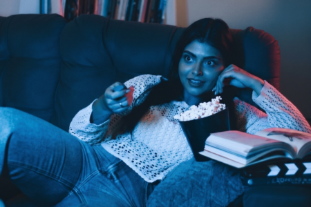 Image: a young woman lies on her sofa watching television, an open book beside her and a bucket of popcorn in front of her.