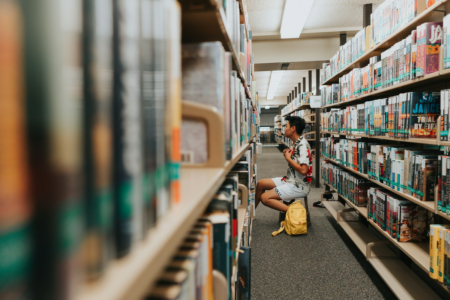 Image: a young man with a backpack sits on a portable stool between two stacks of library shelves, perusing books.