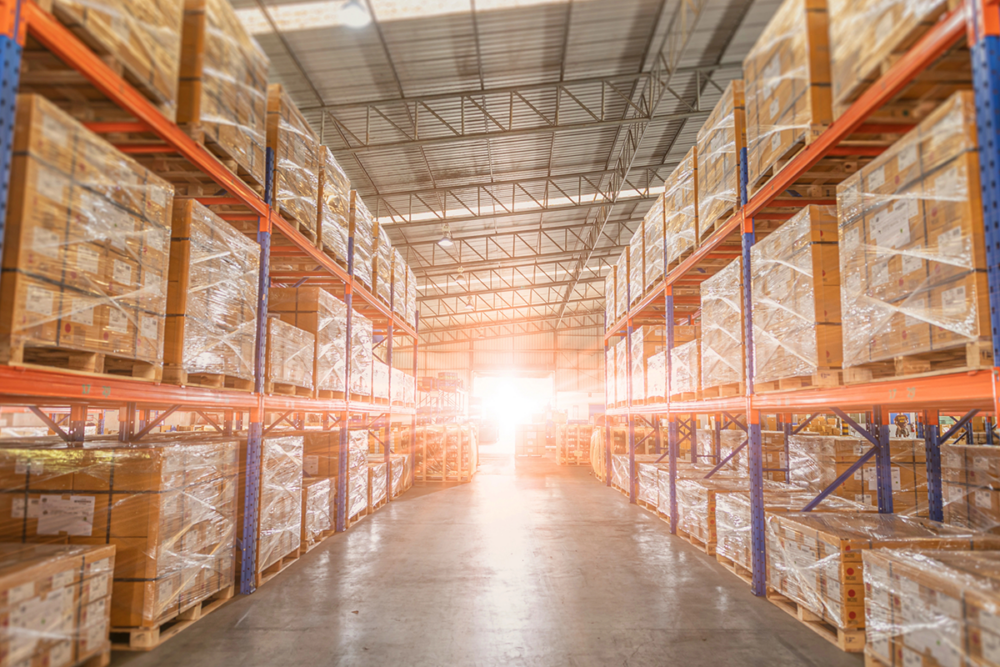 Image: bright dawn light shines in the open door of a warehouse, illuminating the ceiling-high racks full of cartons loaded on shrink-wrapped pallets.