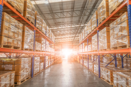 Image: bright dawn light shines in the open door of a warehouse, illuminating the ceiling-high racks full of cartons loaded on shrink-wrapped pallets.