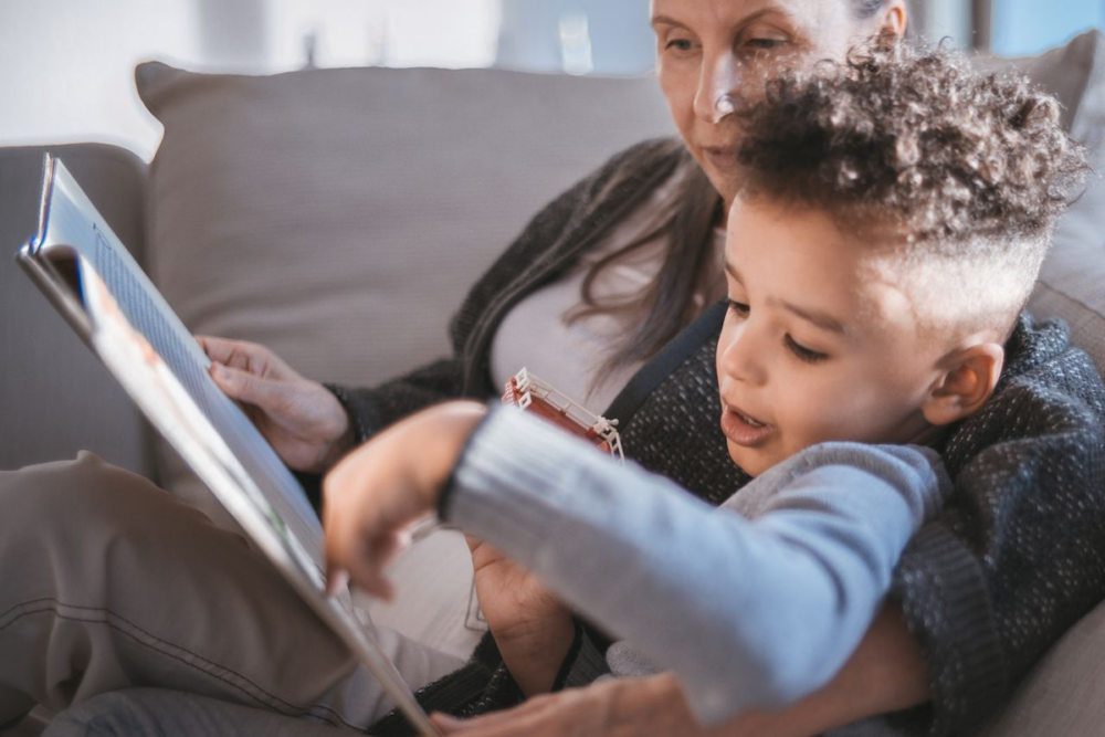 Image: a boy is nestled against his grandmother as they read a picture book together on a sofa.