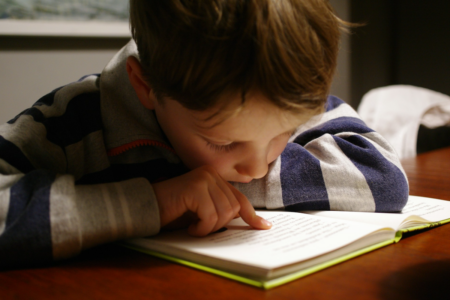 Image: a boy leans closely into the book he's reading, tracing the text with his finger.