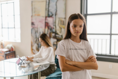 Image: a sullen-looking girl stands with her arms crossed, while in the background her mother works at a laptop computer.