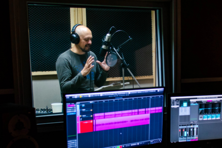 Image: seen through the window of a control booth, a voice actor stands in a recording studio, speaking into a microphone while gesturing expressively with his hands.